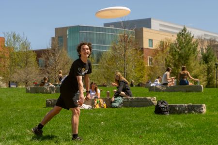 Student plays Frisbee on the campus mall with students talking and studying in the background.