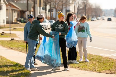 A group of five students holds bags near the side of the road as they pick up trash.