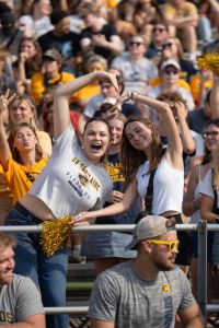 Two students make a heart with their arms with many Blugolds in the background during the Homecoming football game.