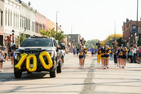 A fun view of the Homecoming parade featuring a car with the number "100" in balloons on the front while people march alongside.