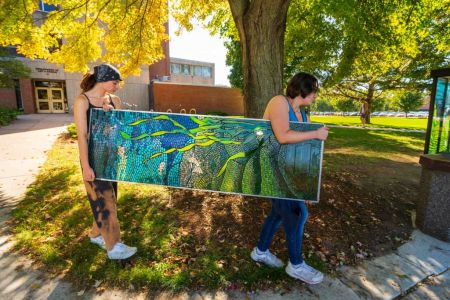 Two students carry a stained glass panel past Hass fine arts. The panel is beautiful, with green and blue, depicting under the Chippewa River.