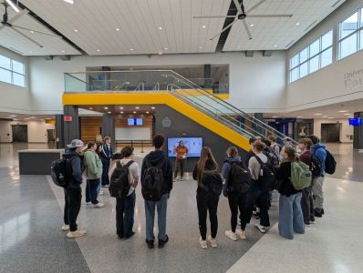 A class of students circles up around a tour lead, who stands in front of the energy dashboard in the lobby of the Sonnentag.