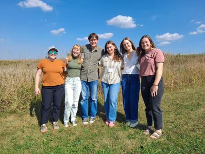 A group of five students stands with their advisor in a prairie, smiling in the sun.