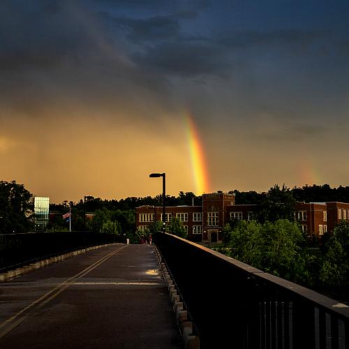 A rainbow emerges from the clouds and appears to end on Schofield Hall as seen from the footbridge.