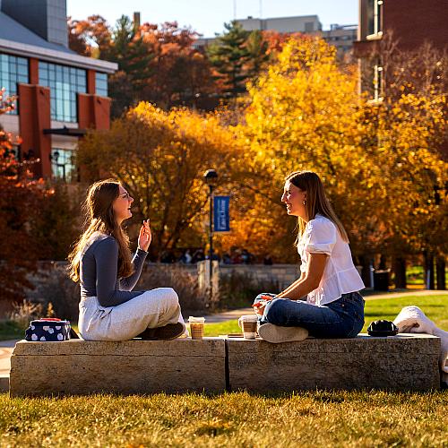 Two students sit on a rock on campus mall, laughing, talking, and drinking coffee