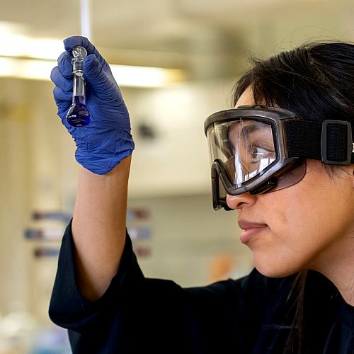 A student wearing safety goggles closely examines liquid in a small test tube