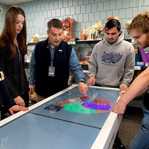 A group of students listen as a professor talks and points at a digital table with an image of a brain on it.