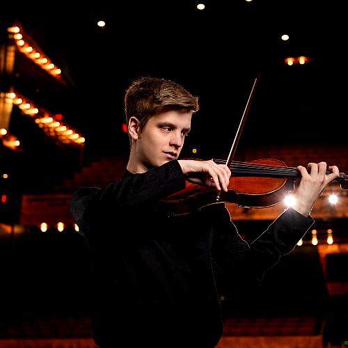 A student practices a violin solo in a dimly lit rehearsal hall. 