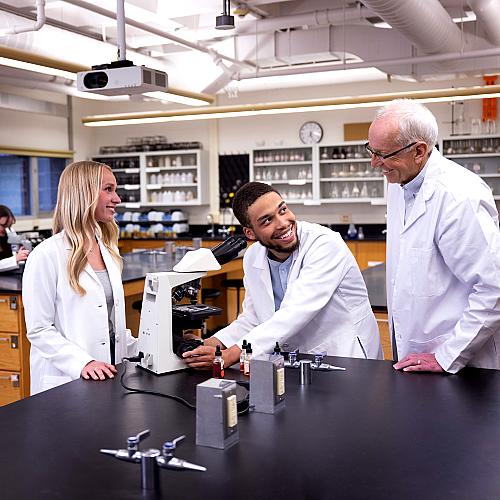 Students and a professor in white lab coats work together in a lab, smiling.