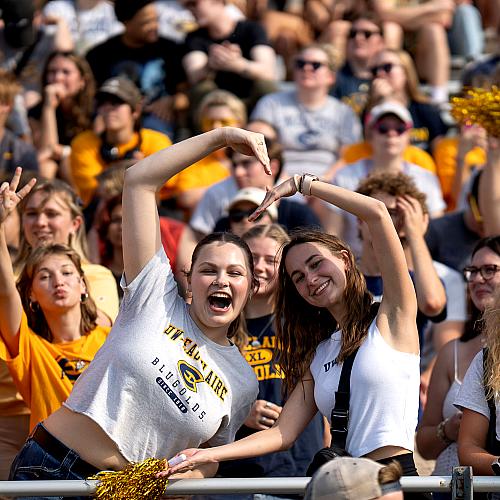 Two students make a heart with their arms with many Blugolds in the background during the Homecoming football game.