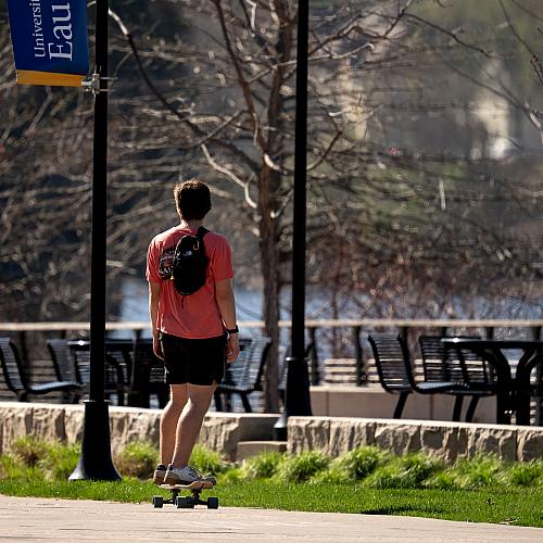 A student wearing shorts, a t-shirt, and a backpack skateboarding on the campus sidewalk